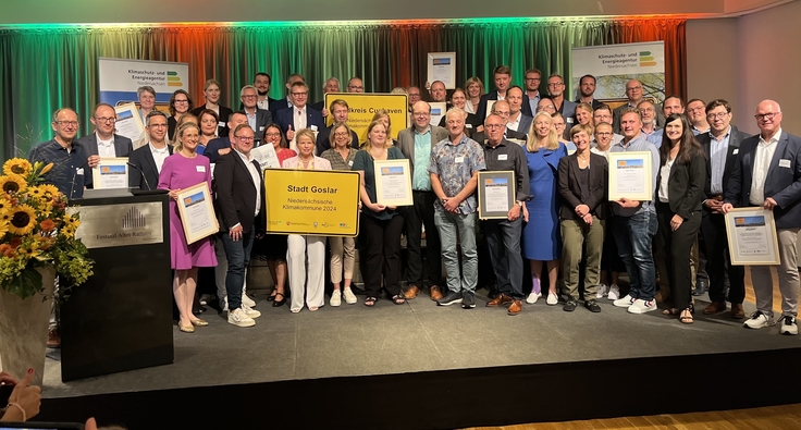 Gruppenfoto der Preisträgerinnen und Preisträger des niedersächsischen Wettbewerbs „Klima kommunal 2024“ auf einer Bühne mit Urkunden in der Hand.