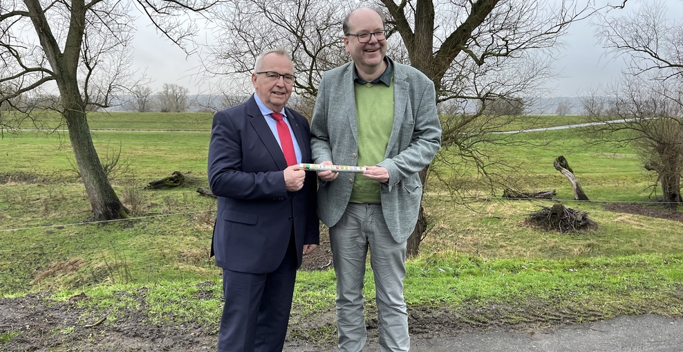 Minister Meyer bekommt vor einer grünen Wiese den symbolischen Staffelstab von Kollege Backhaus aus M-V überreicht.